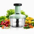 A countertop electric food chopper with a mint green body and black handle, surrounded by fresh vegetables including lettuce, bell peppers, carrots, onions, and peas on a white background.